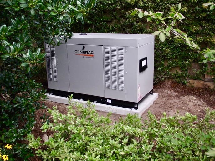 Large grey Generac standby generator installed on a concrete pad surrounded by lush green bushes.